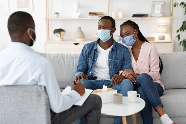 Family Therapy During Pandemic. Happy Black Couple In Protective Masks Sitting On Couch At Counselor's Office, Listening Advices, Selective Focus